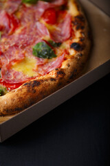 Pizza with cheese, ham, green basil leaves and tomatoes in paper box on black background. Food product advertising banner, copy space