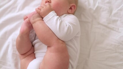 Portrait baby in white onesie lies on soft bed, lifting its legs and grabbing them with tiny hands, infant appears playful, body coordination, baby growth, home comfort, childhood curiosity