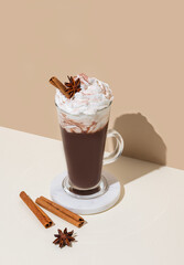 Hot chocolate with whipped cream, cinnamon and spices in a tall glass on a white and beige background