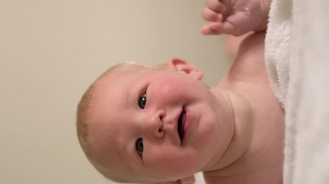 Vertical footage, smiling infant baby lies on their tummy, looking up with bright eyes, Cild face expresses pure joy and curiosity, baby emotions, childhood happiness, home comfort