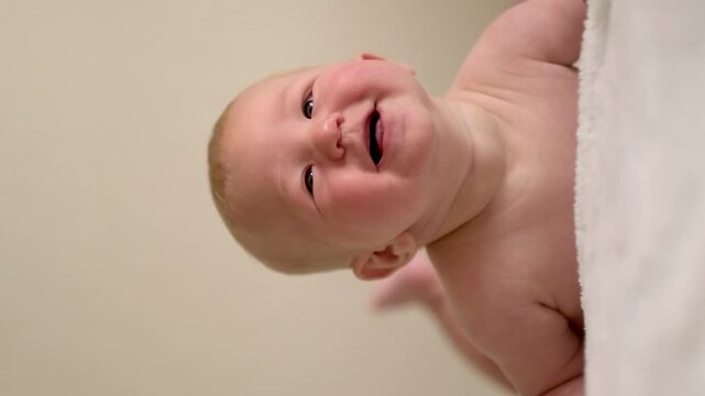 Vertical footage, smiling infant baby lies on their tummy, looking up with bright eyes, Cild face expresses pure joy and curiosity, baby emotions, childhood happiness, home comfort