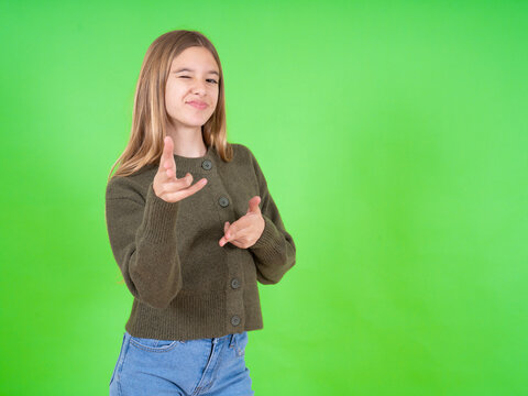 Joyful beautiful teen girl wearing green sweater wink and points index fingers at camera, chooses someone, makes finger gun pistol.