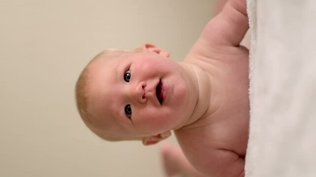 Vertical footage, smiling infant baby lies on their tummy, looking up with bright eyes, Cild face expresses pure joy and curiosity, baby emotions, childhood happiness, home comfort