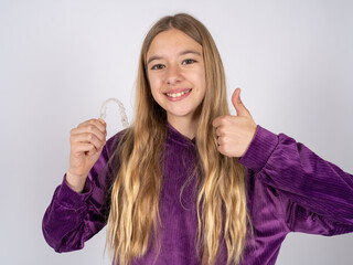 beautiful teen girl wearing purple sweater holding an invisible braces aligner and rising thumb up, recommending this new treatment. Dental healthcare concept.