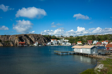 Obraz premium Hamburgsund, Sweden, a scenic coastal village with traditional white and red houses by the water. Wooden piers stretch over the calm bay, with rocky cliffs in the background under a blue sky