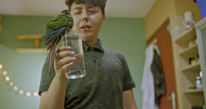 A young girl holds a glass of water for her pet green parrot to drink. The parrot perches on the glass and drinks. They are at home.