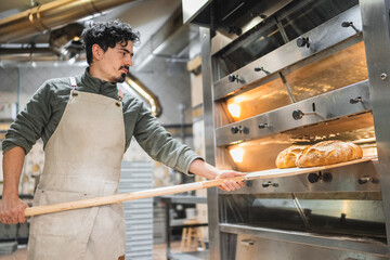 Baker taking freshly baked bread out of oven in bakery