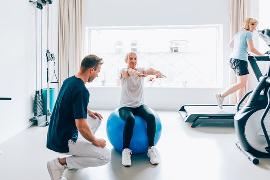 Elderly woman performing balance exercises on a stability ball under supervision of a male trainer in a bright, modern gym setting