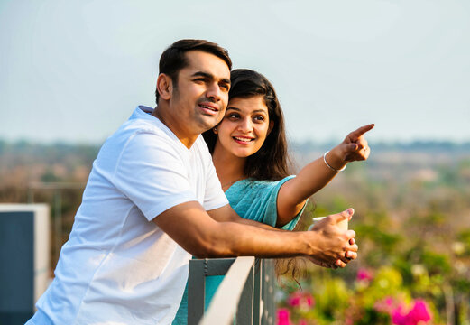 Young Indian couple sharing joyful moments on a balcony, chatting, pointing, and sipping coffee