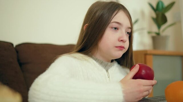 Girl taking big bite of red apple while sitting indoors. Female enjoying fresh fruit. Lady savoring crisp apple in cozy home setting. Child eating healthy snack with enthusiasm
