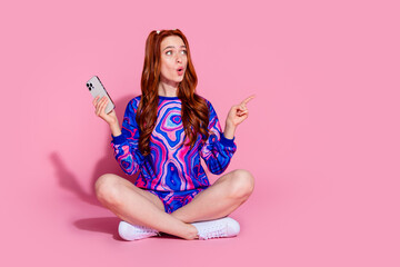 Full size photo of young girl sit floor hold device point look empty space wear trendy blue print outfit isolated on pink color background