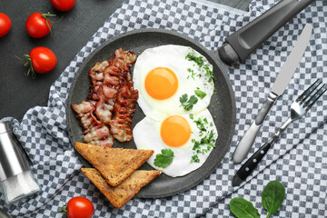 Tasty fried eggs, bacon and pieces of toast served on black table, flat lay