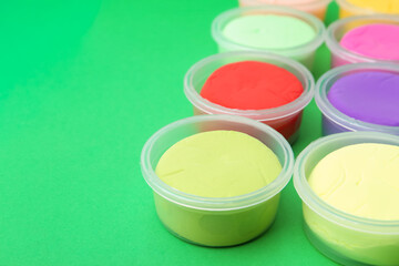 Different colorful modeling clay in plastic jars on green background, closeup. Space for text