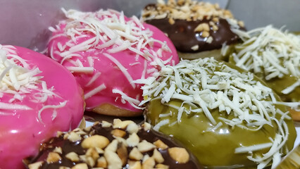 Close-up photo of various donuts in a box. The top is topped with colorful sprinkles, cheese and nuts, with the focus on the front of the doughnut.