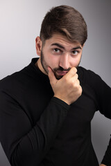 portrait d'un beau jeune homme de 25 ans. Il est brun, barbu et souriant et il porte un pull over noir. Arrière plan gris