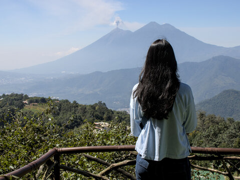 woman with brown hair looking at an erupting volcano in antigua guatemala (vulcan de fuego and acatenango mountains in the background) hiking travel tourism adventure getaway latin central america