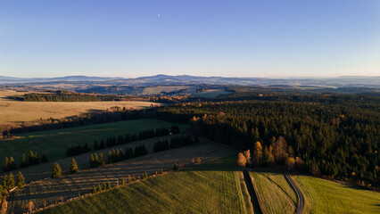 Fototapeta premium Aerial view of a vast countryside at sunset, with rolling hills, dense forests, and open fields. The golden light casts long shadows, highlighting the beauty of the landscape. Orlické hory, Czechia.