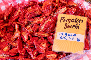 Close-up of sun-dried tomatoes with deep red tones and rough uneven surfaces piled in a plastic-lined bin with a label indicating origin and price at an Italian market
