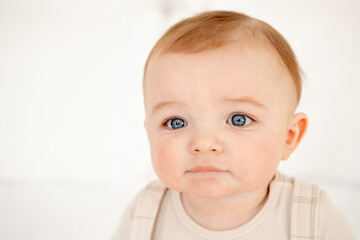 A baby on a white bed is smiling, a cute portrait of a small child a boy with blue eyes in beige clothes is sitting on a bed in a bedroom at home, a smiling baby