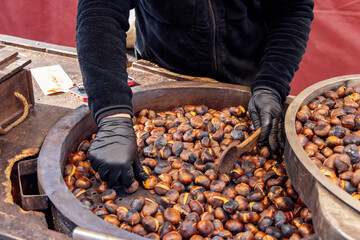 Street vendor roasting chestnuts in a large metal pan gloved hands stir the hot nuts with a wooden spoon as they develop a golden brown and charred texture