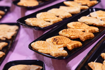 Freshly baked taiyaki fish-shaped pastries with a golden crispy crust are arranged on black trays served as a traditional Japanese street food dessert with various sweet fillings