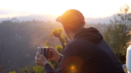 Male hiker wearing orange cap savoring hot beverage at sunrise, overlooking expansive mountain...