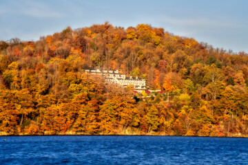 Obraz premium a stunning autumn landscape with a hillside blanketed by trees in full fall colors—orange, red, and yellow. A serene blue lake lies in the foreground, reflecting the surrounding foliage.