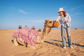 Female tourist petting a saddled camel in a desert landscape, United Arab Emirates