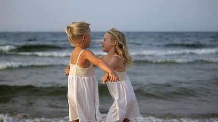 Blonde twin sisters sharing warm ocean embrace, wearing matching white dresses while standing barefoot along serene shoreline during golden hour sunset