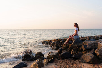 woman sits on the rocky beach, reflecting on life and savoring the beautiful colors that light up...