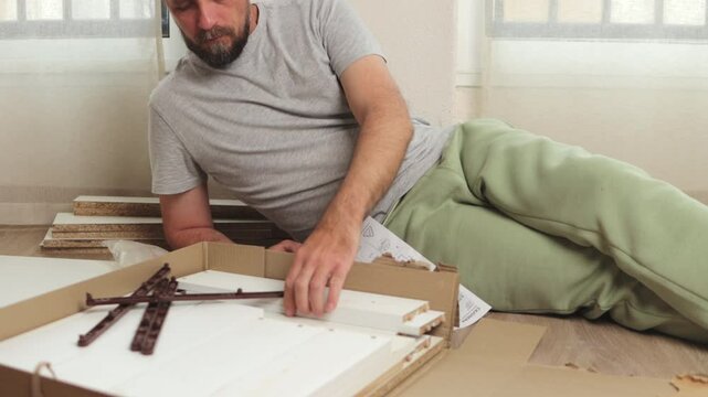 Furniture assembly process involves a man carefully reading instructions and holding a screwdriver, sitting amidst various furniture parts and tools in his home