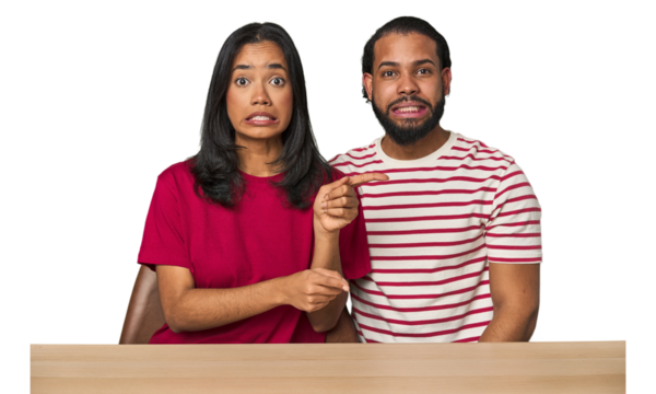Seated young Latino couple at table shocked pointing with index fingers to a copy space.