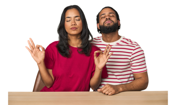 Seated young Latino couple at table relaxes after hard working day, she is performing yoga.