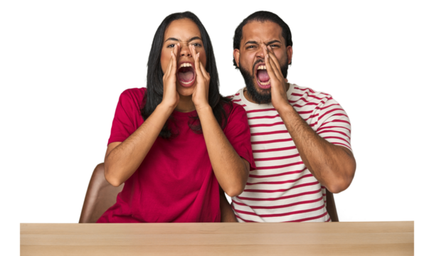Seated young Latino couple at table shouting excited to front.