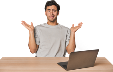 Hispanic young man working on laptop confused and doubtful shrugging shoulders to hold a copy space.