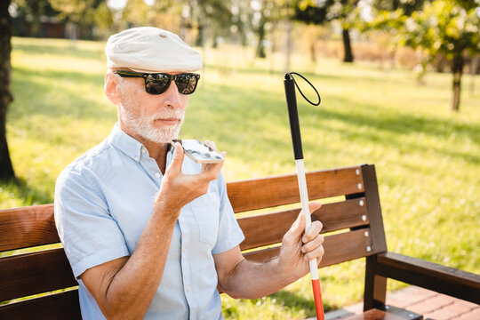 Elderly gray-haired caucasian entrepreneur with low vision in sunglasses sitting on bench, having a phone call using speaker mode, and holding white walking crane with one hand. Outdoor closeup shot