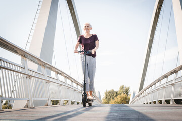 one woman mature senior caucasian female standing on electric kick scooter in day riding driving in...