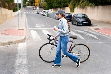 Young woman walking with bicycle and coffee across crosswalk