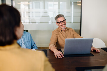 Smiling businessman wearing glasses and a brown shirt, sitting at a conference table with a laptop....