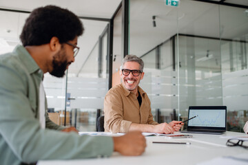 Fototapeta premium Smiling businessman leading a corporate presentation, discussing financial growth with colleagues in a modern office. Engaged team members in productive discussion, fostering collaboration and success