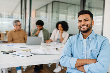 Fototapeta premium A young businessman in a casual denim shirt sits confidently at a modern workspace, exuding positivity while colleagues engage in a team discussion.
