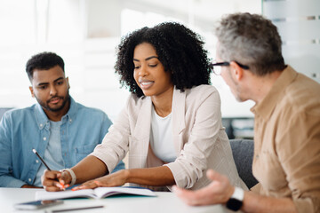 A diverse business team in a modern office discussing a project, smiling and engaging around a laptop. Professional collaboration, teamwork, and strategy planning in a dynamic corporate environment.