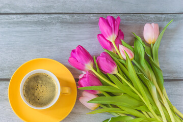 Spring coffee break concept: yellow cup of coffee and bouquet of purple and pink tulips on a wooden table; top view