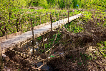 Crooked footbridge over a ditch with a stream, plastic trash in the ditch