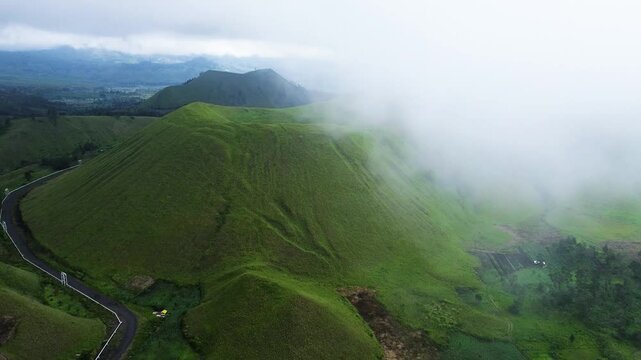 aerial footage of towering green hills surrounded by green meadows enveloped in a mystical fog. view of green hills peak with grassland and mountain views of Wurung Crater, Indonesia