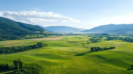 Fototapeta premium Rolling green hills and meadows stretch across valley between mountain ranges, sunlit agricultural landscape with scattered forests under blue summer sky.
