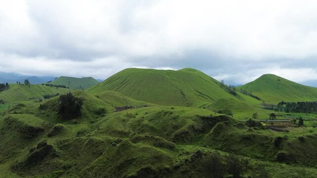Stunning aerial footage of Kawah Wurung, Indonesia, showcasing towering green hills surrounded by expansive lush grasslands and green meadows.