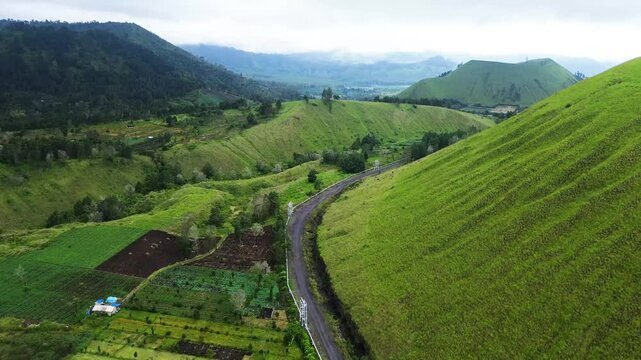 Stunning aerial footage tracking winding roads through vast green hills and geothermal power pipelines, surrounded by misty grasslands in Wurung Crater, Indonesia.