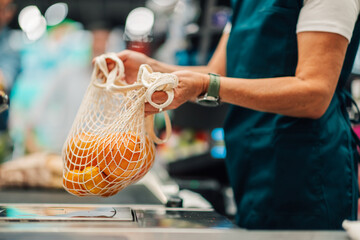 Cashier weighing oranges in reusable bag at supermarket checkout