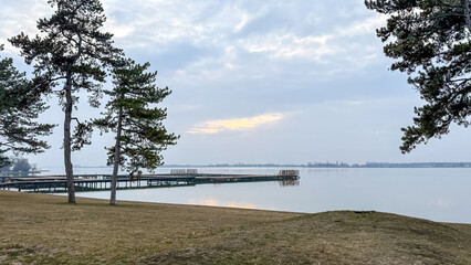 Serene lakeside view with pine trees and pier under cloudy sky, nature relaxation, peaceful...